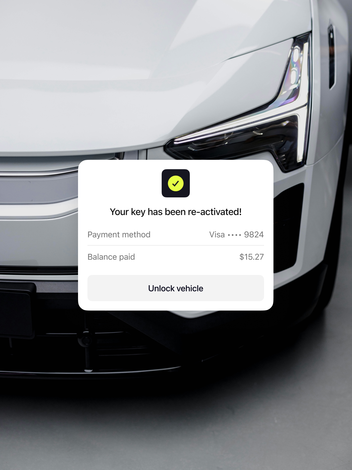 A close-up of a white car's front with a digital message on the screen, stating "Your key has been re-activated!" The message indicates payment made using a Visa card ending in 9824 and shows a balance of $15.27, with an "Unlock vehicle" button.