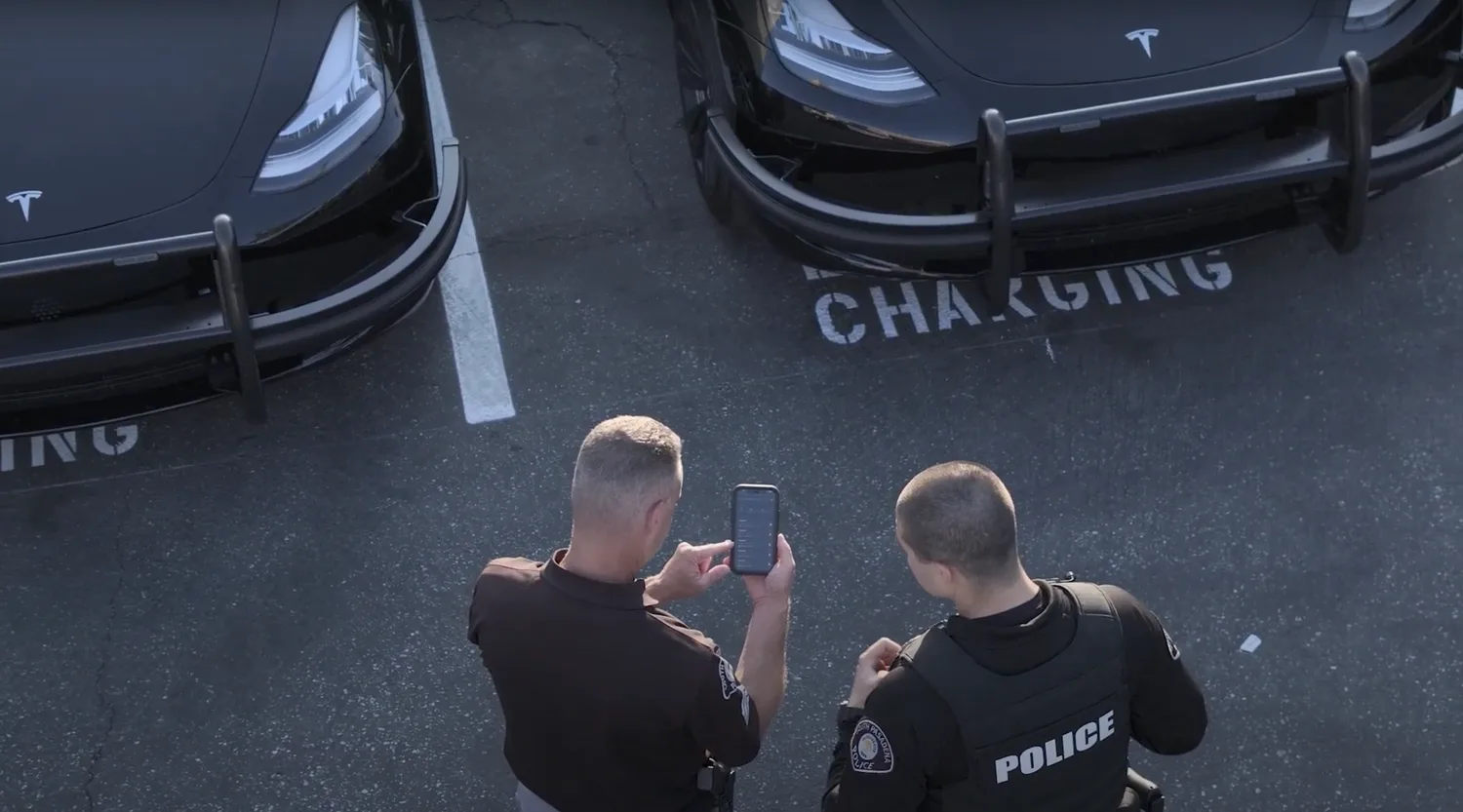 Two individuals, one in a police uniform, are discussing near parked black Tesla vehicles in a charging station. The officer is checking information on a smartphone.