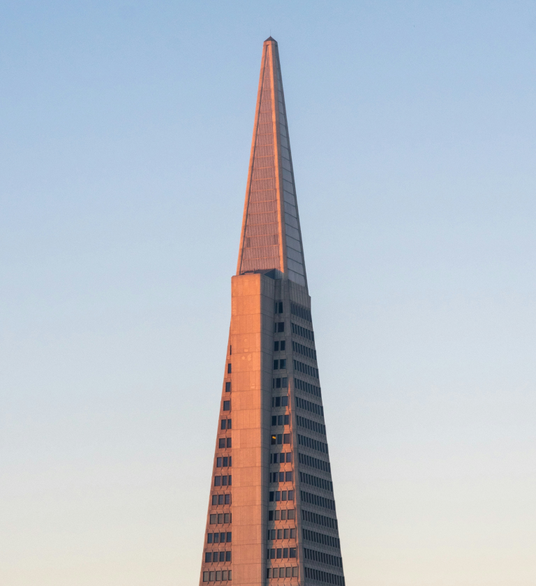 The top of a modern triangular skyscraper featuring a pointed spire, against a clear blue sky during sunset.