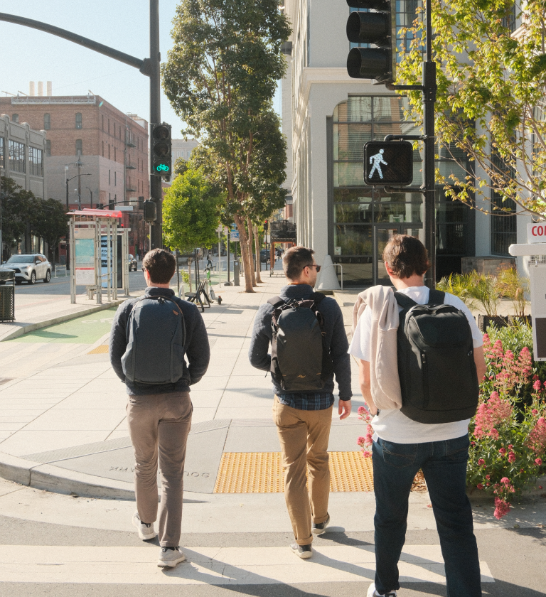 Three individuals walking on a city sidewalk, each with a backpack. They are heading towards a crosswalk with a pedestrian signal visible. Behind them, urban buildings and greenery are present.