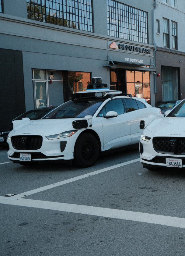 Two white electric vehicles parked at a crosswalk in an urban area. The vehicle on the left has a roof-mounted sensor. In the background, a building with large windows and a sign visible.
