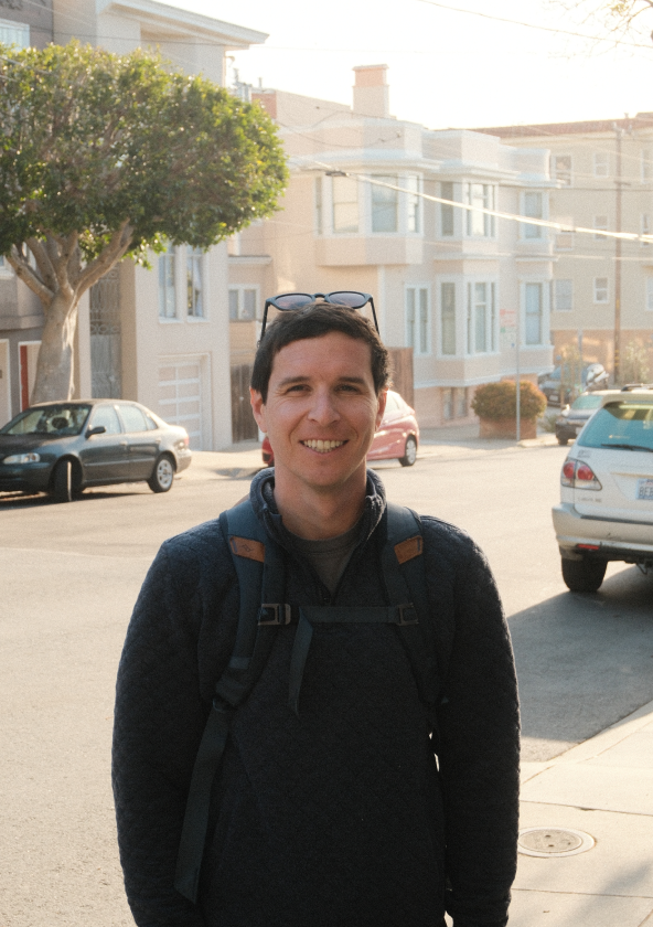 A man standing on a street with a smile, wearing a dark sweater and a backpack. In the background, there are residential buildings, parked cars, and trees, indicating a suburban area.