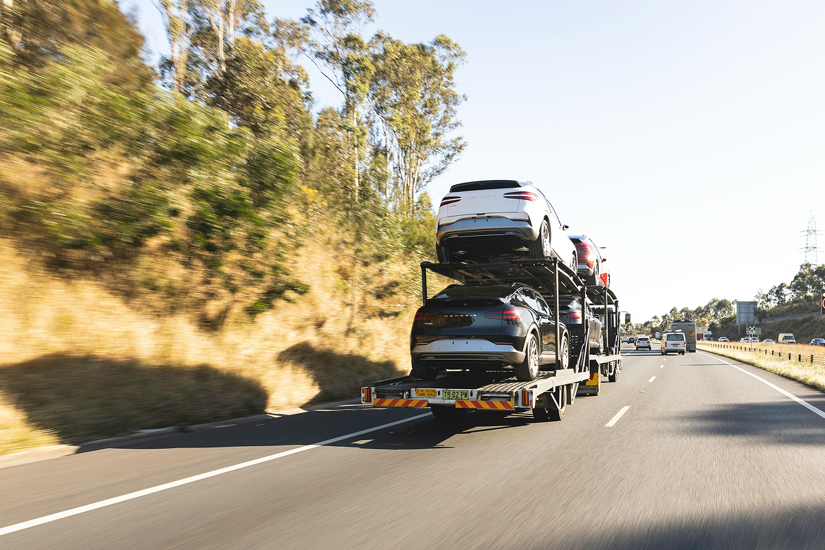 A transport truck carrying multiple cars stacked on a trailer, driving on a highway surrounded by trees and under a clear blue sky.