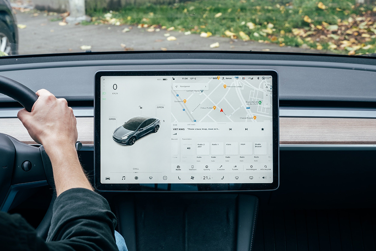 A driver using a touchscreen display in a car, showing a digital map, vehicle status, and media options. The dashboard includes a large screen with various app icons and navigation features.
