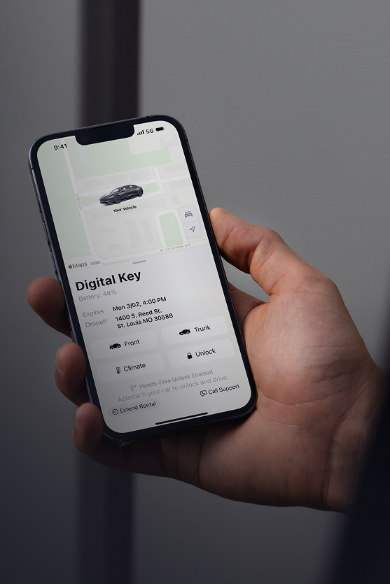 A hand holding a smartphone displaying a digital key app with a car image and options for unlocking, climate control, and trunk access. The background shows a blurred indoor setting.